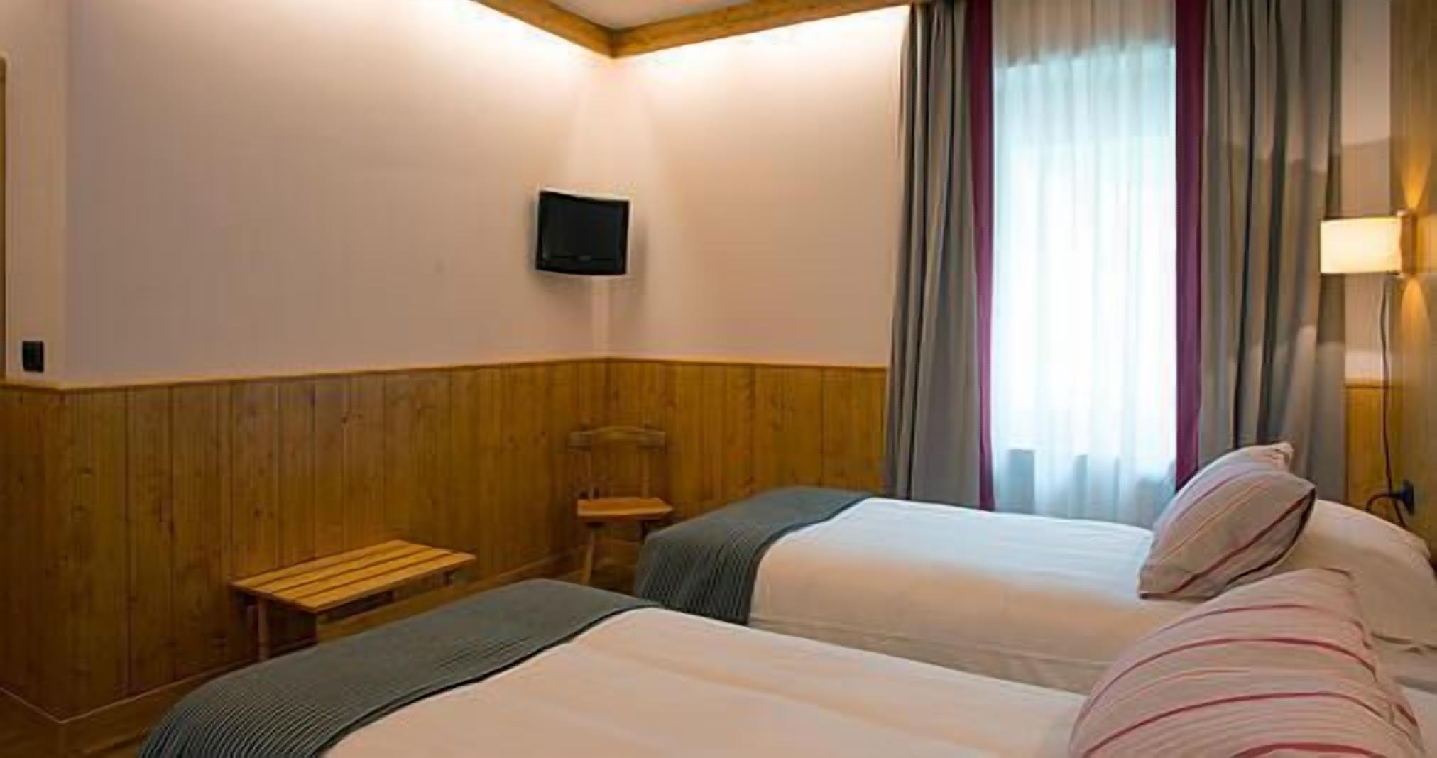 Une chambre d’hôtel avec deux lits, un petit banc en bois, un téléviseur fixé au mur et une fenêtre avec des rideaux.