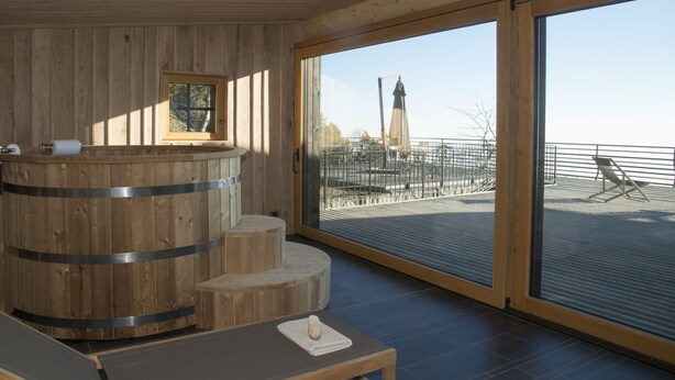 Ein hölzerner Spa-Bereich mit Whirlpool, Sitzgelegenheiten und einem großen Fenster mit Blick auf einen entfernten Turm.