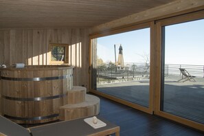 Een houten wellnessruimte met een jacuzzi, zitplaatsen en een groot raam met uitzicht op een verre toren.