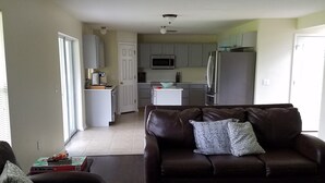 A living room with a brown leather sofa, a patterned throw pillow, and a small table. A kitchen with white cabinets and a microwave is visible in the background.