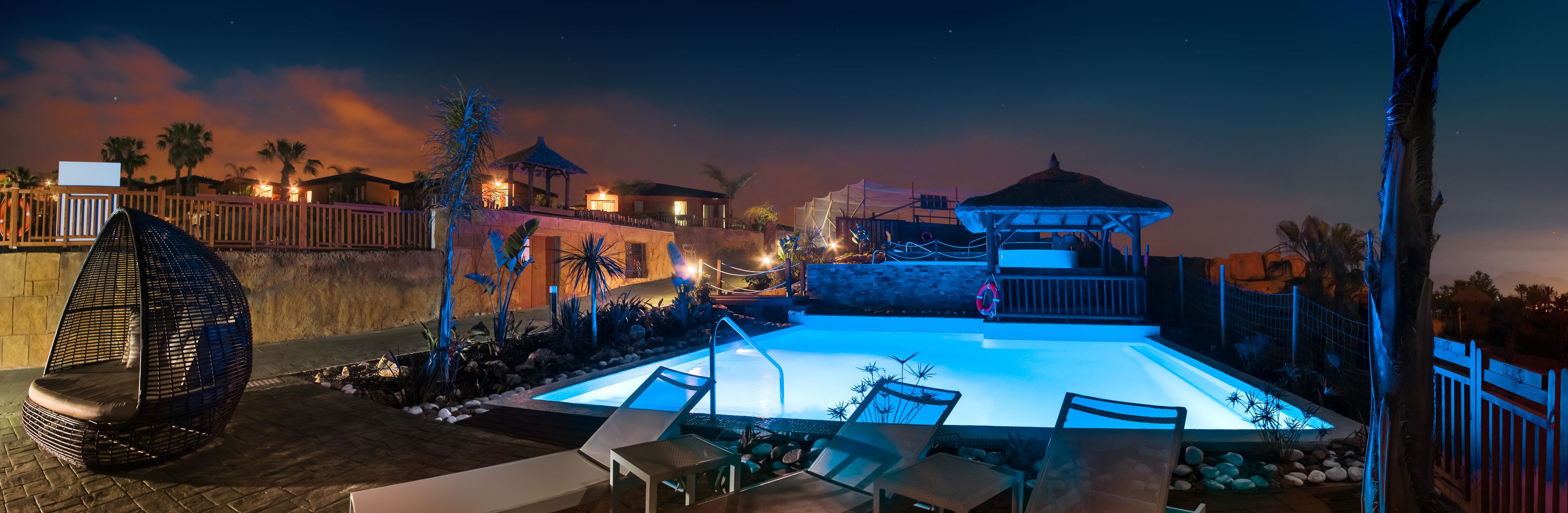 Vista nocturna de un área de piscina con sillones reclinables, fuente de agua y un pabellón.