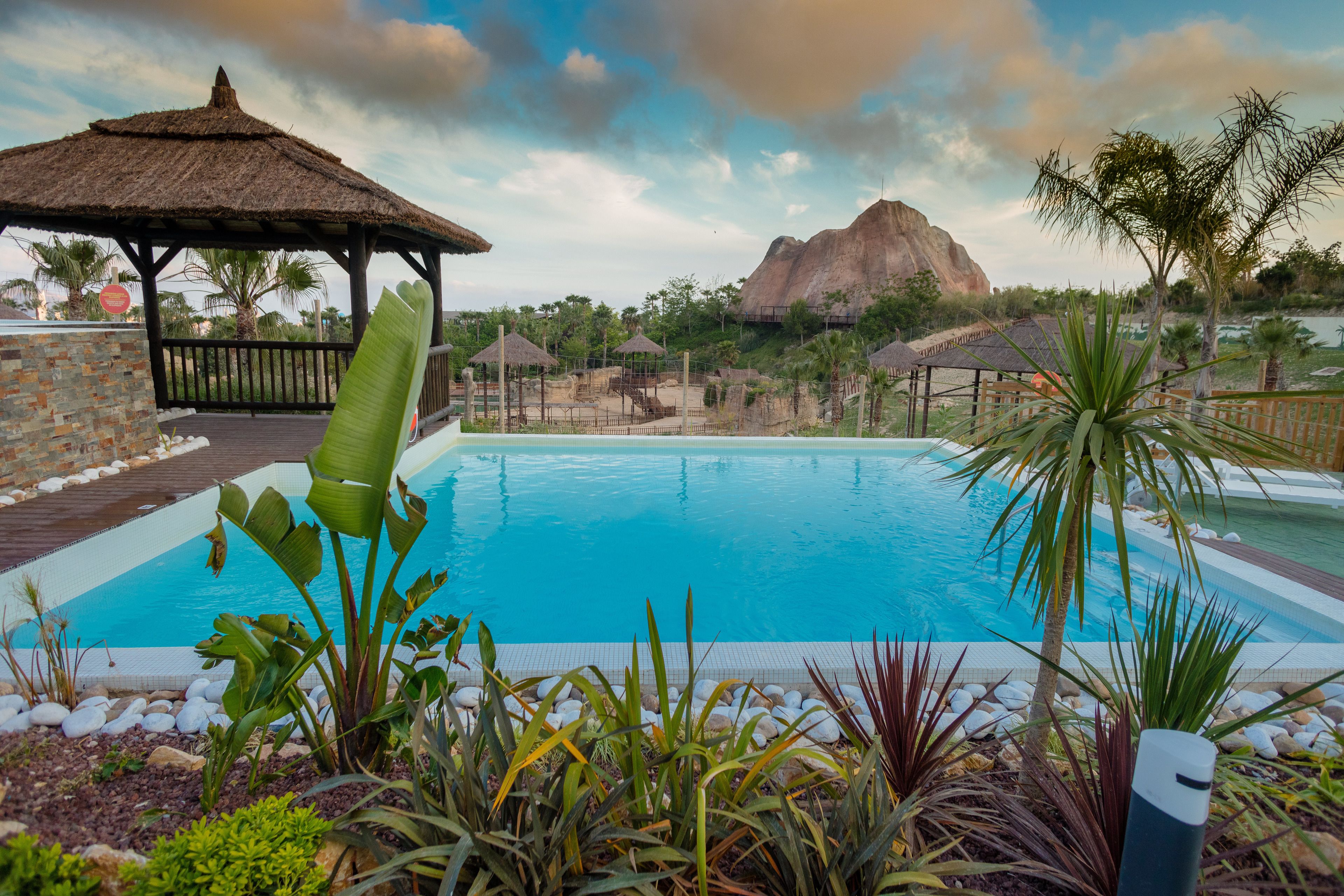 Una piscina con un pabellón de techo de paja, rodeada de plantas tropicales y montañas al fondo.