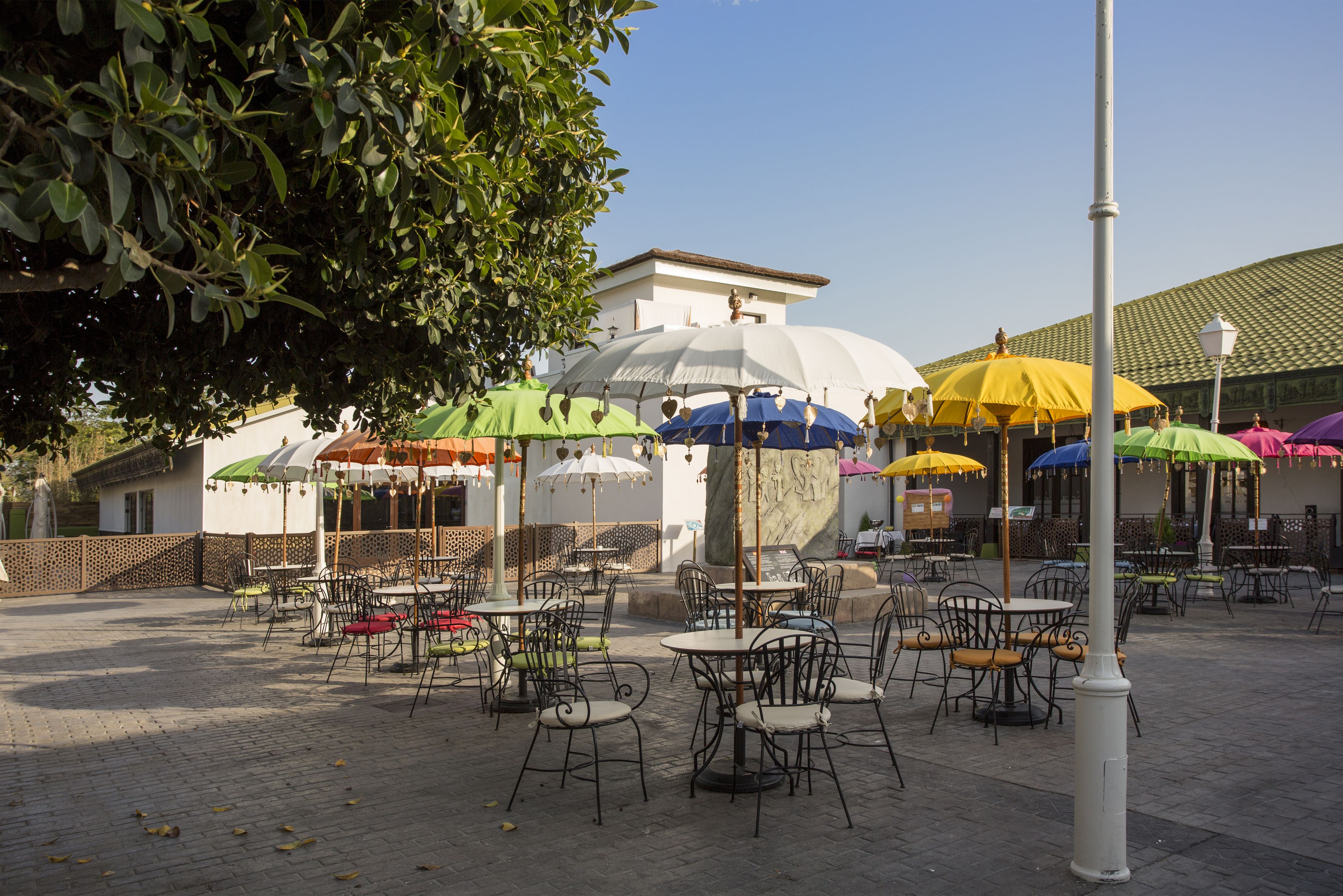 Un espace de café en plein air avec des tables, des chaises, des parasols et un bâtiment en arrière-plan.