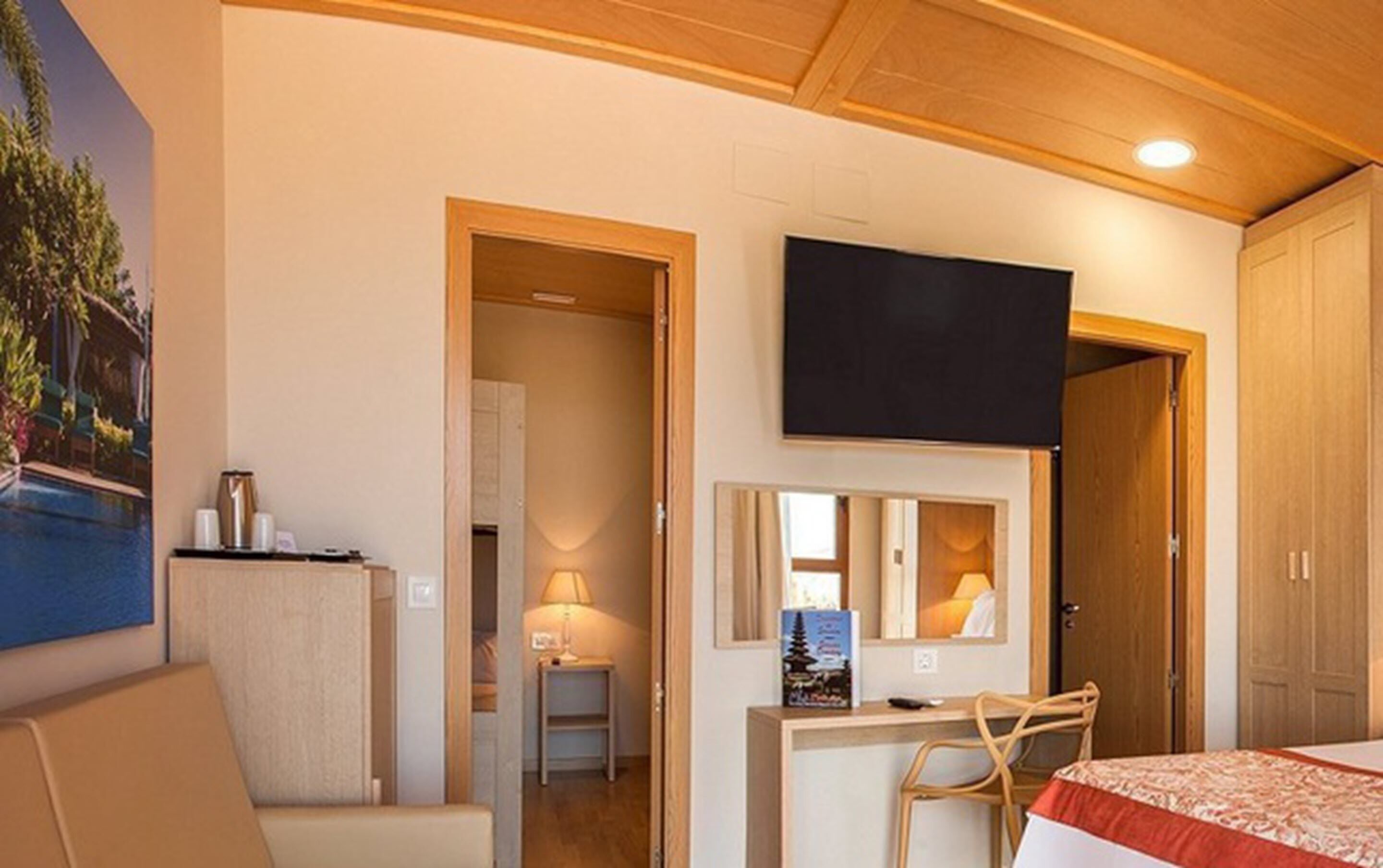 A hotel room with a bed, a desk with a chair, a television mounted above the desk, and a painting on the wall.