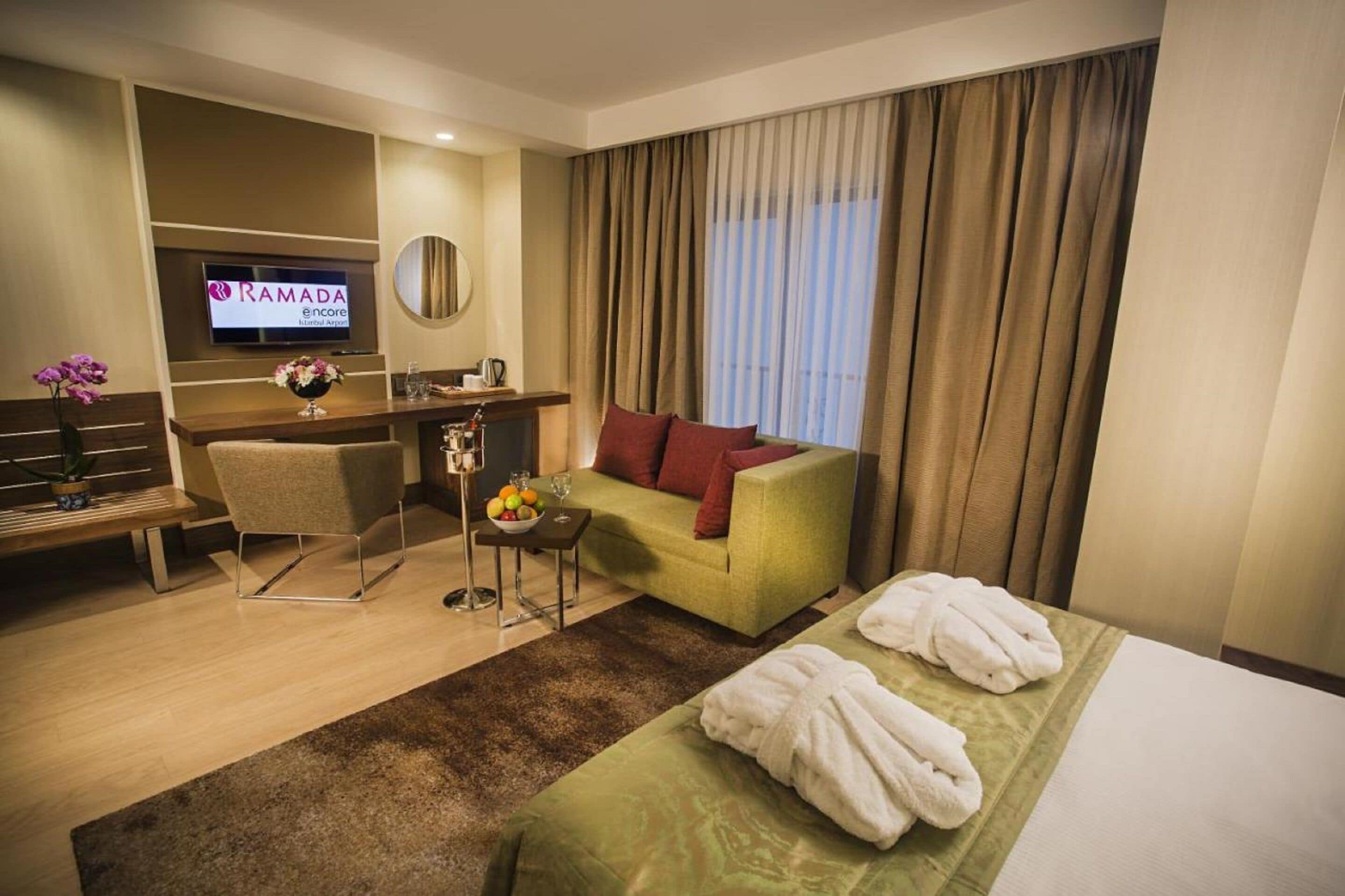 Ein Hotelzimmer mit einem Bett, einem grünen Sofa, einem Schreibtisch mit Fernseher, einem Sessel und einem kleinen Tisch mit Obstschale und Gläsern.