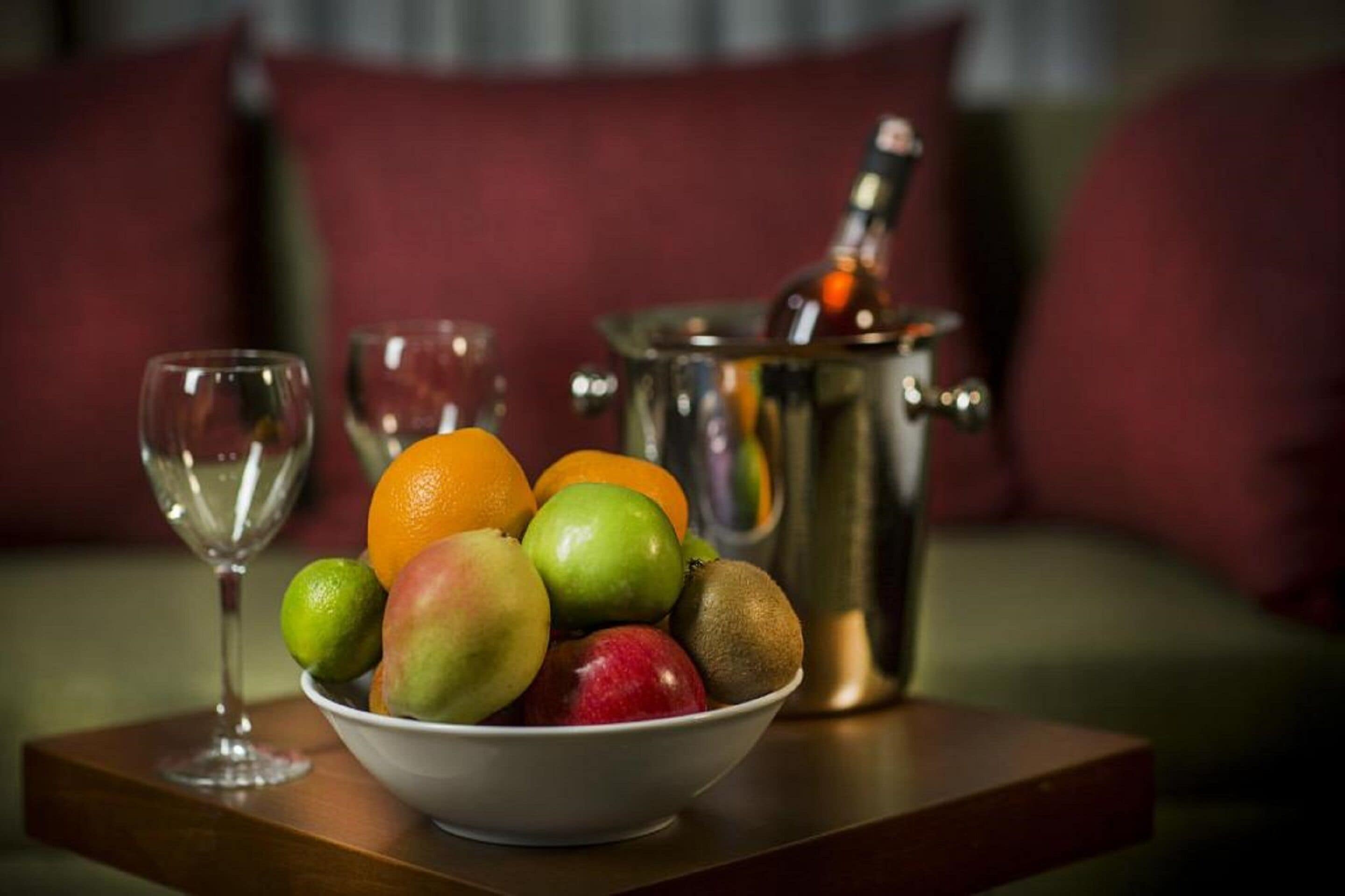 A bowl of fruit on a wooden tray with a wine glass and a champagne bottle in the background.