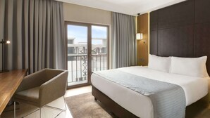 A hotel room with a large bed, a desk, and a chair. There is a balcony with a view of buildings.