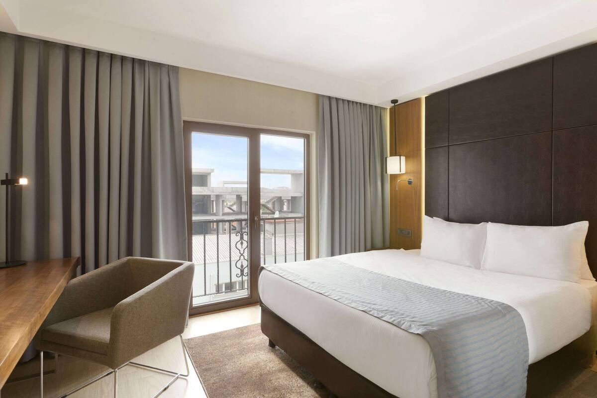 A hotel room with a large bed, a desk, and a chair. There is a balcony with a view of buildings.