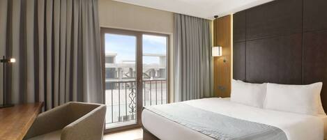 A hotel room with a large bed, a desk, and a chair. There is a balcony with a view of buildings.