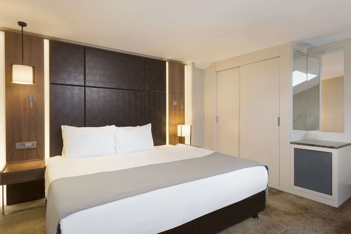 A modern hotel room with a large bed, a nightstand, a wall-mounted lamp, and a wardrobe with a mirror.