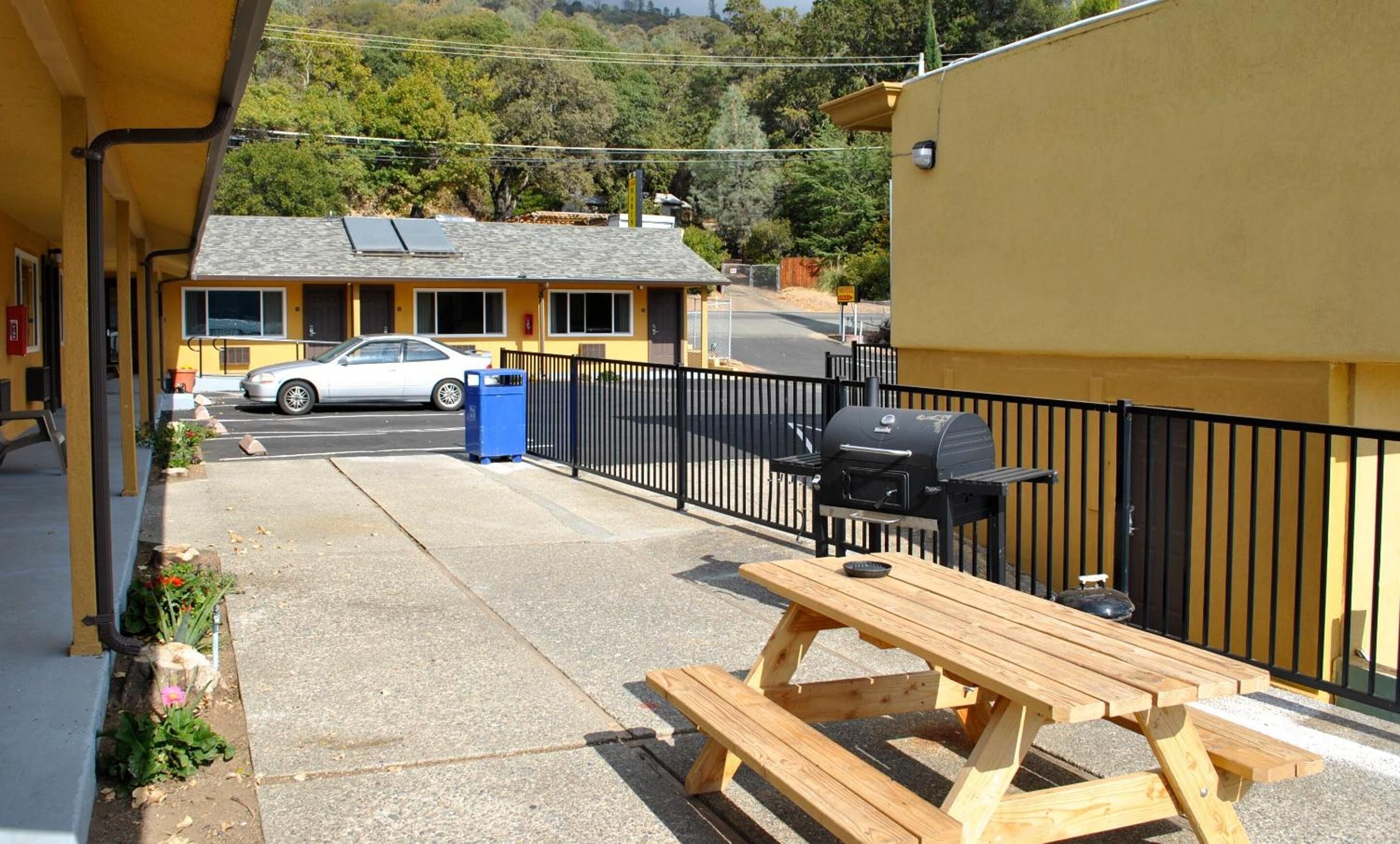 Un patio con una mesa y una parrilla de madera, un coche estacionado frente a un edificio amarillo, y vistas a una colina boscosa.