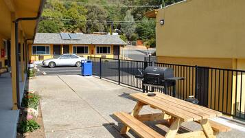 Un patio con una mesa y una parrilla de madera, un coche estacionado frente a un edificio amarillo, y vistas a una colina boscosa.