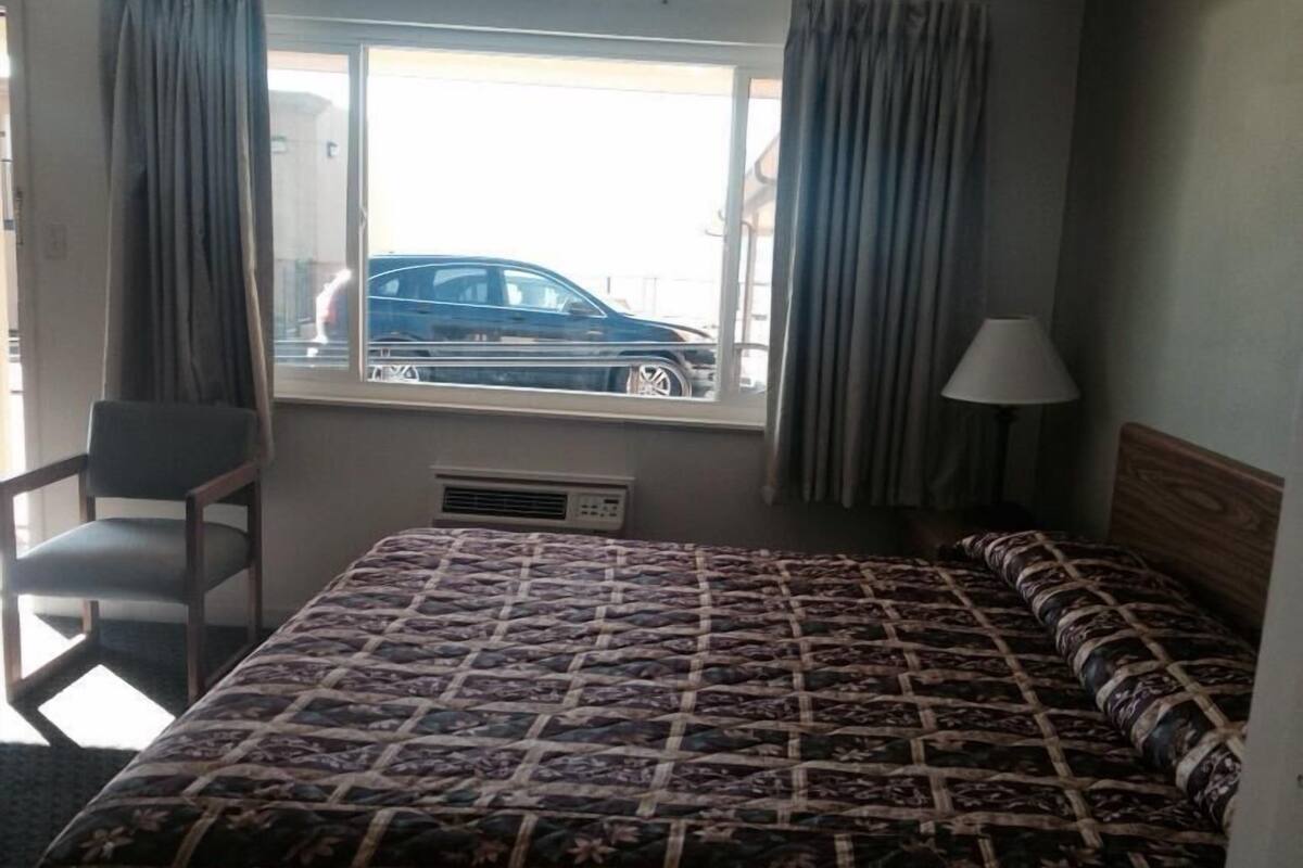 A hotel room with a bed, a chair, a window with curtains, and a lamp.