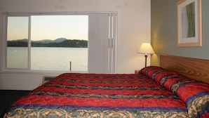 A hotel room with a large bed, a window with a view of a lake and mountains, a bedside lamp, and a framed picture on the wall. - Lake Marina Inn (Clearlake Oaks)
