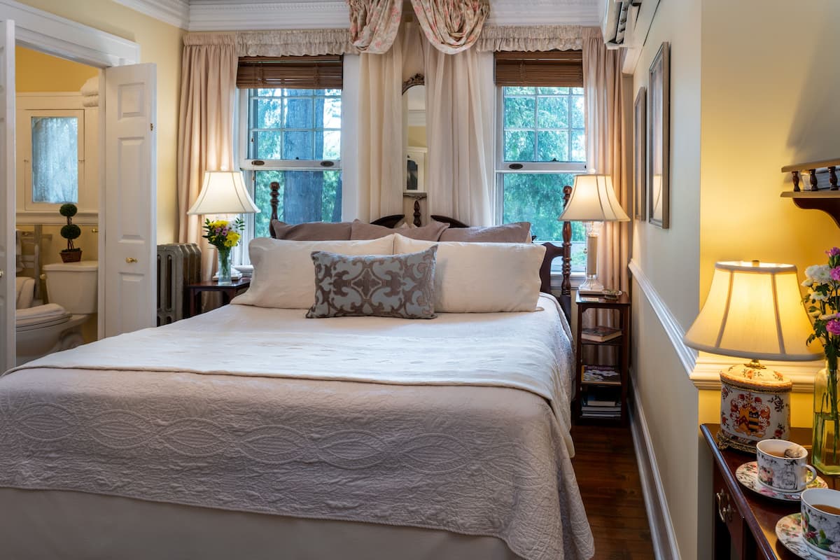 Une chambre à coucher avec un grand lit, deux tables de chevet avec des lampes, un miroir au-dessus du lit et une fenêtre avec des rideaux.