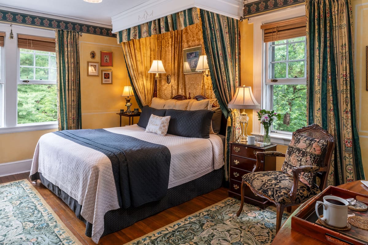 Une chambre à coucher avec un lit à baldaquin, deux lampes de chevet, un fauteuil à motifs floraux et une vue sur la verdure par les fenêtres.