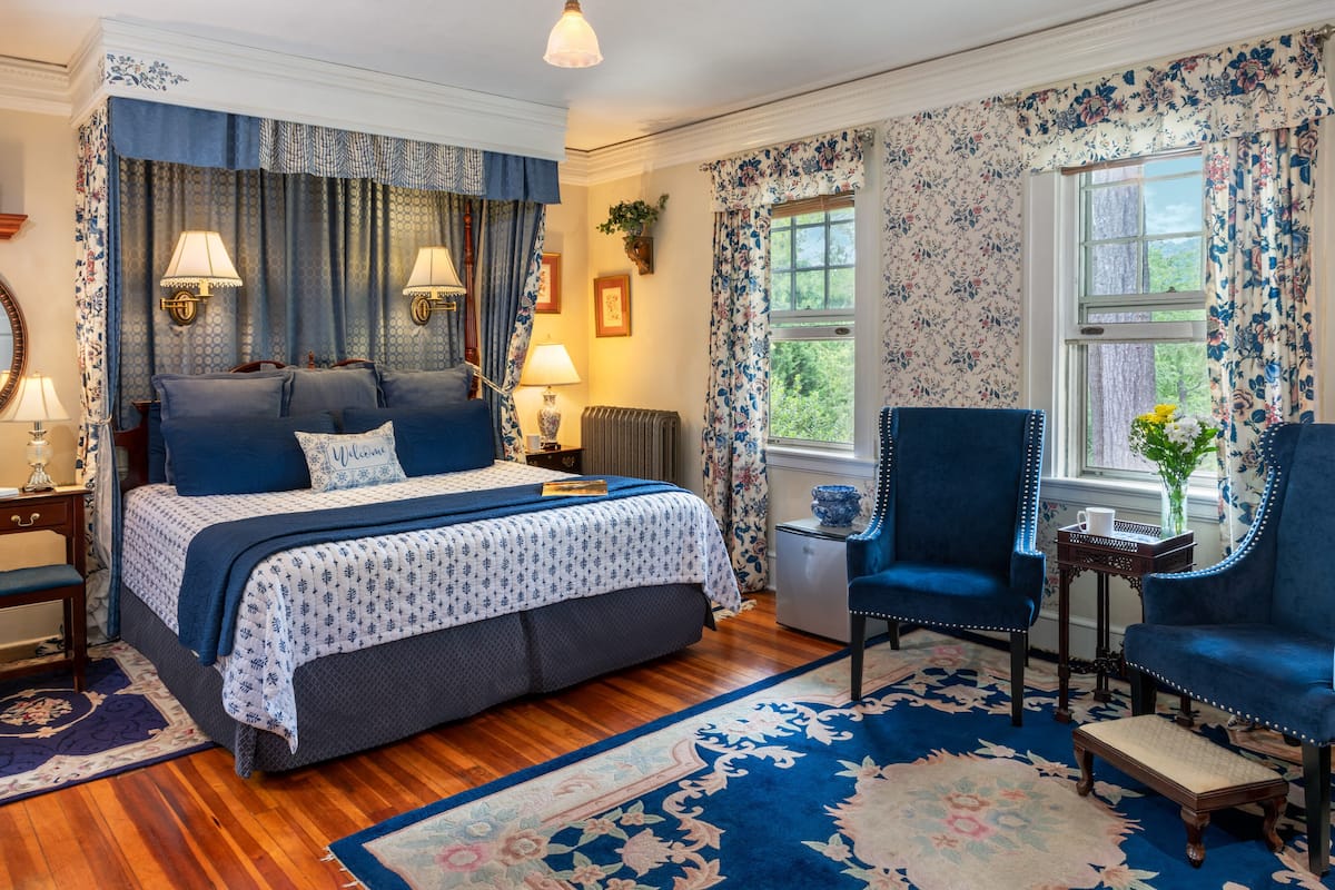 Une chambre à coucher avec un grand lit, deux chaises, une petite table et une fenêtre ornée de rideaux à motifs floraux.