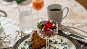 A table setting with a glass dessert bowl containing fruit and granola, a glass of orange juice, a cup of coffee, and a slice of bread on a patterned plate.
