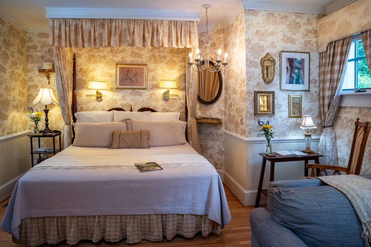 Une chambre à coucher classique, avec un grand lit, deux tables de chevet avec des lampes, un lustre et des œuvres d’art encadrées accrochées aux murs.