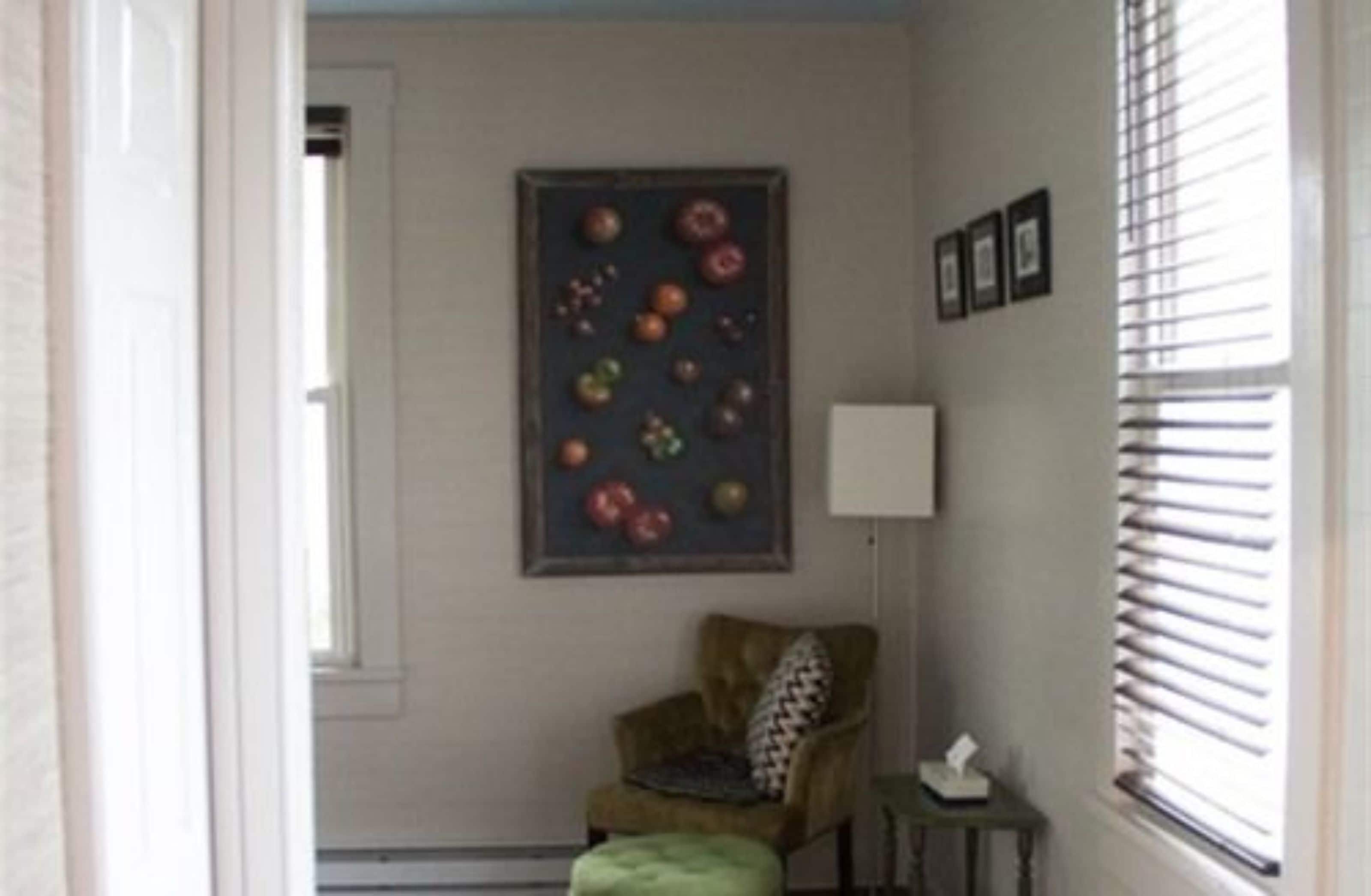 Ein Zimmer mit Tür, einem Bilderrahmen mit Obst, einem Stuhl, einem kleinen Tisch und einem Fenster mit Jalousien.