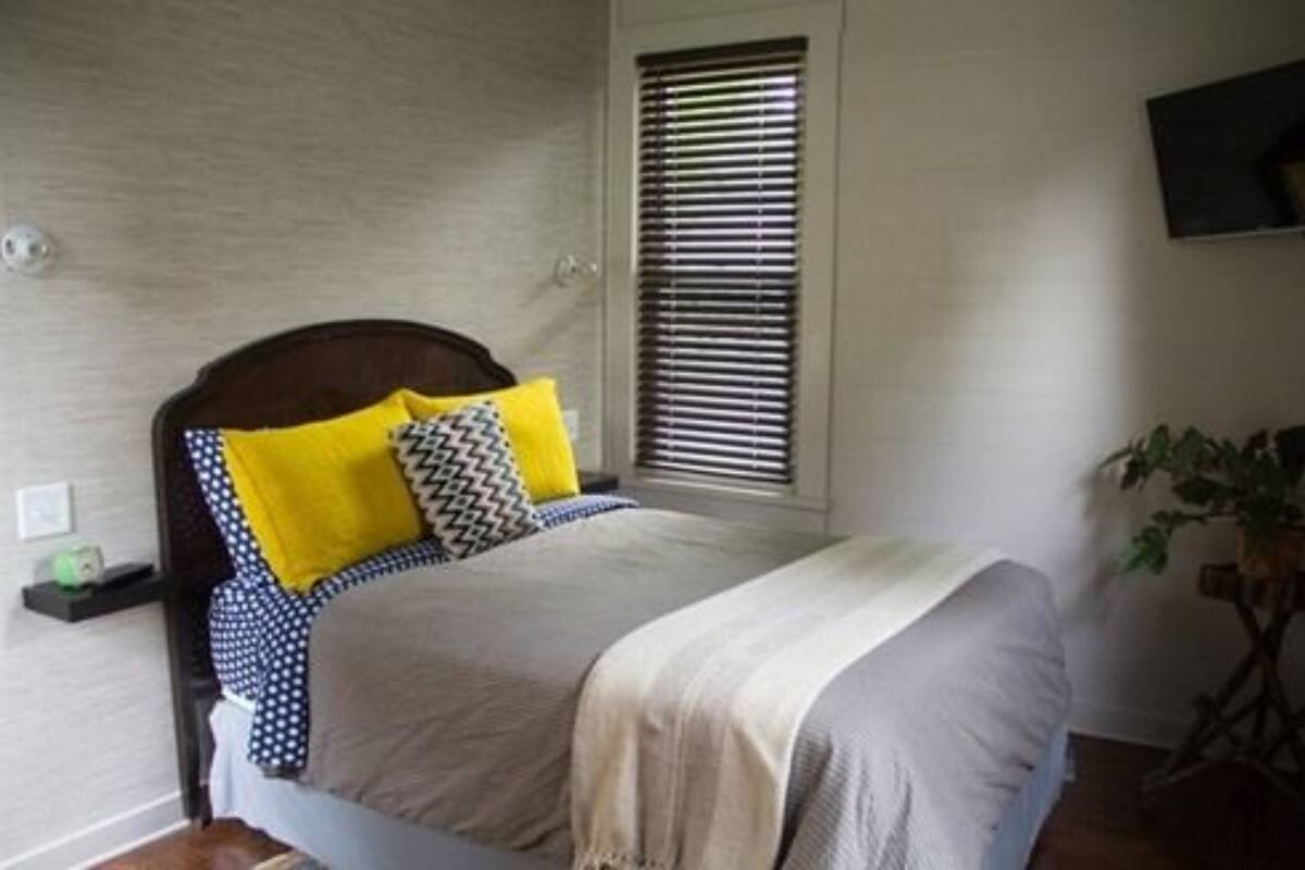 A bedroom with a bed, a window with blinds, a bedside table, and a wall-mounted TV.