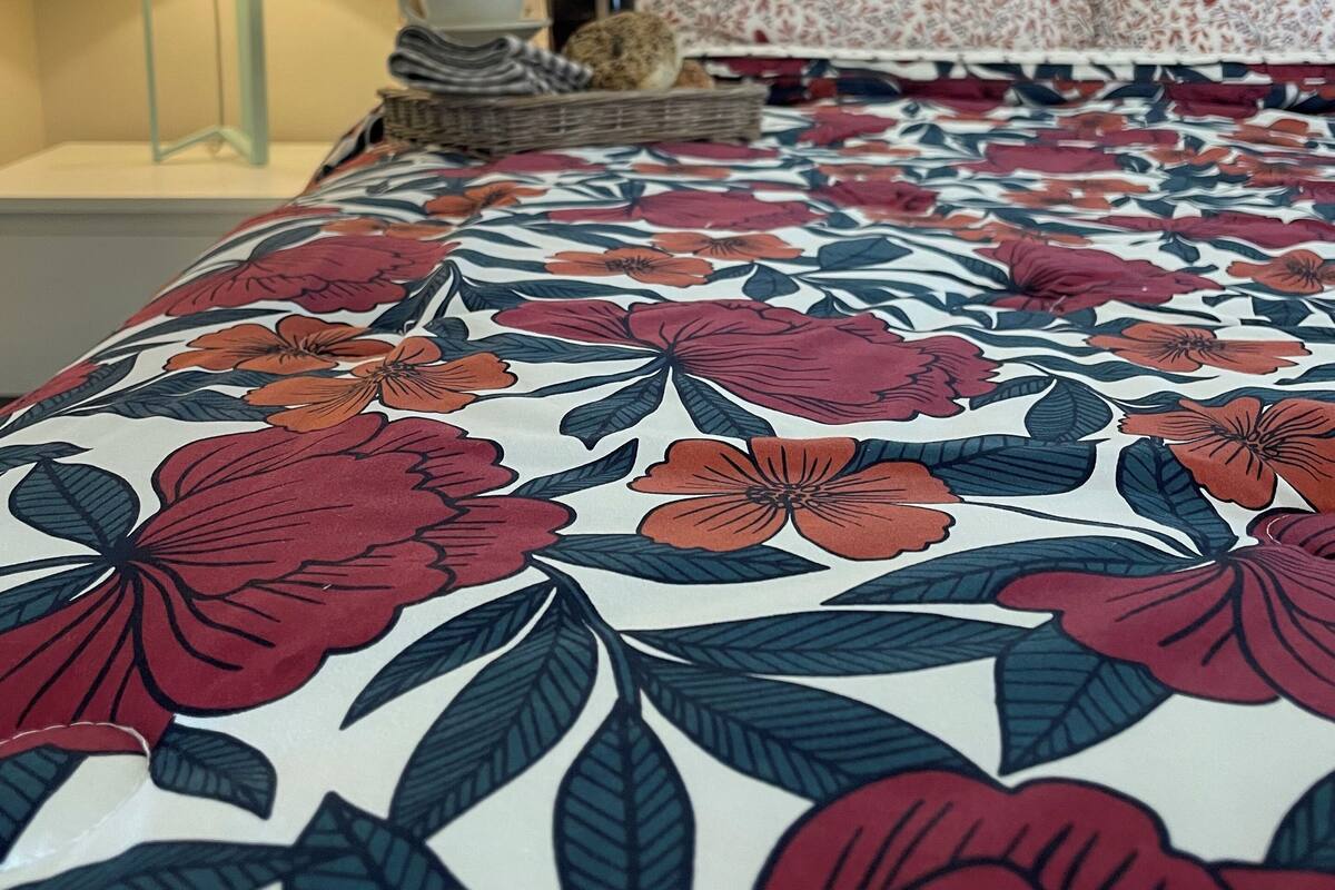 A neatly made bed with a floral-patterned bedspread, a bedside table with a lamp, and a window with blinds.