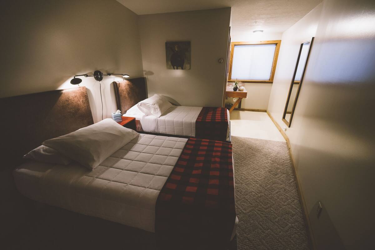 A hotel room with two beds, a nightstand, a lamp, a painting on the wall, and a window.