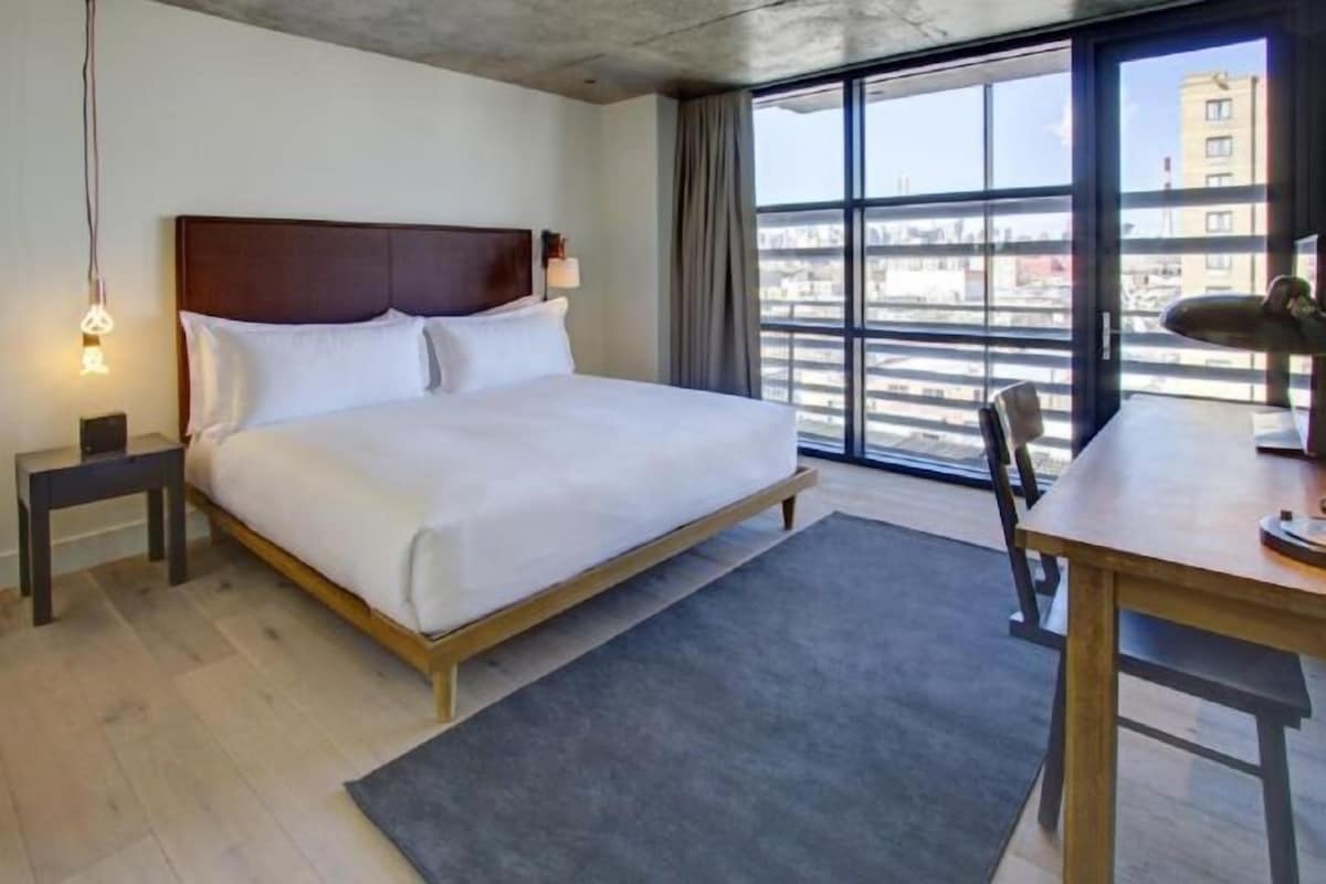 A hotel room with a bed, a desk, a chair, a nightstand, and a large window offering a city view.