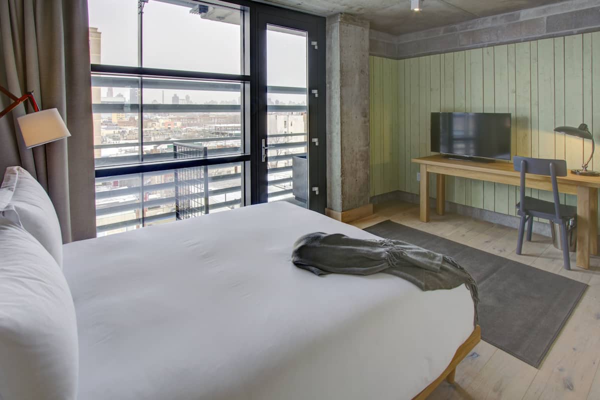 Hotellihuone, jossa on suuri sänky, työpöytä ja televisio sekä ikkunasta avautuva näkymä kaupunkiin.