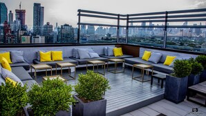 Un lounge en la azotea con vista al horizonte urbano, que cuenta con un conjunto de sofás modernos con cojines amarillos y grises, mesitas y plantas en macetas.