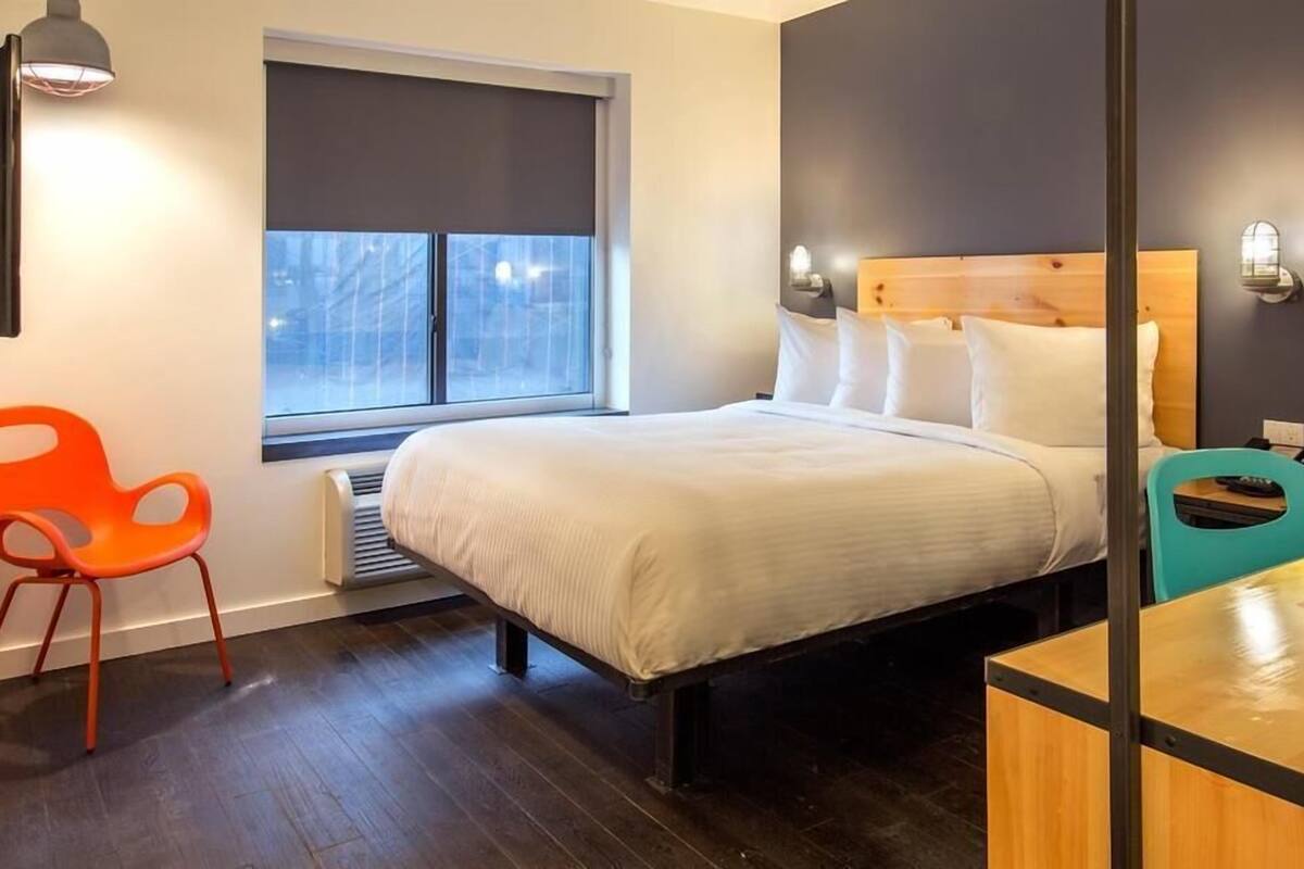 A modern hotel room with a large bed, a window with blinds, a heater, an orange chair, a teal chair, and a wooden nightstand.