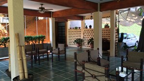 Uma área de estar coberta ao ar livre com cadeiras, plantas em vasos e um ventilador de teto.