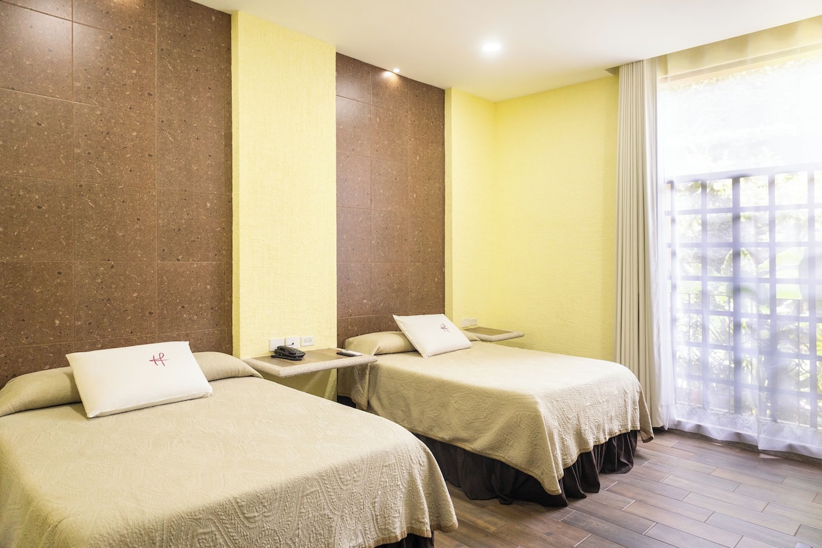 A hotel room with two beds, a small table, and a window with a view.