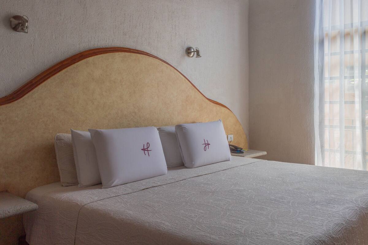 A neatly made bed with a headboard, two pillows with a monogram, and a bedside table with a lamp.