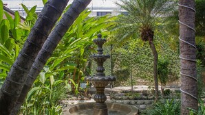 Um jardim com uma fonte central rodeada por diversas plantas e árvores tropicais.
