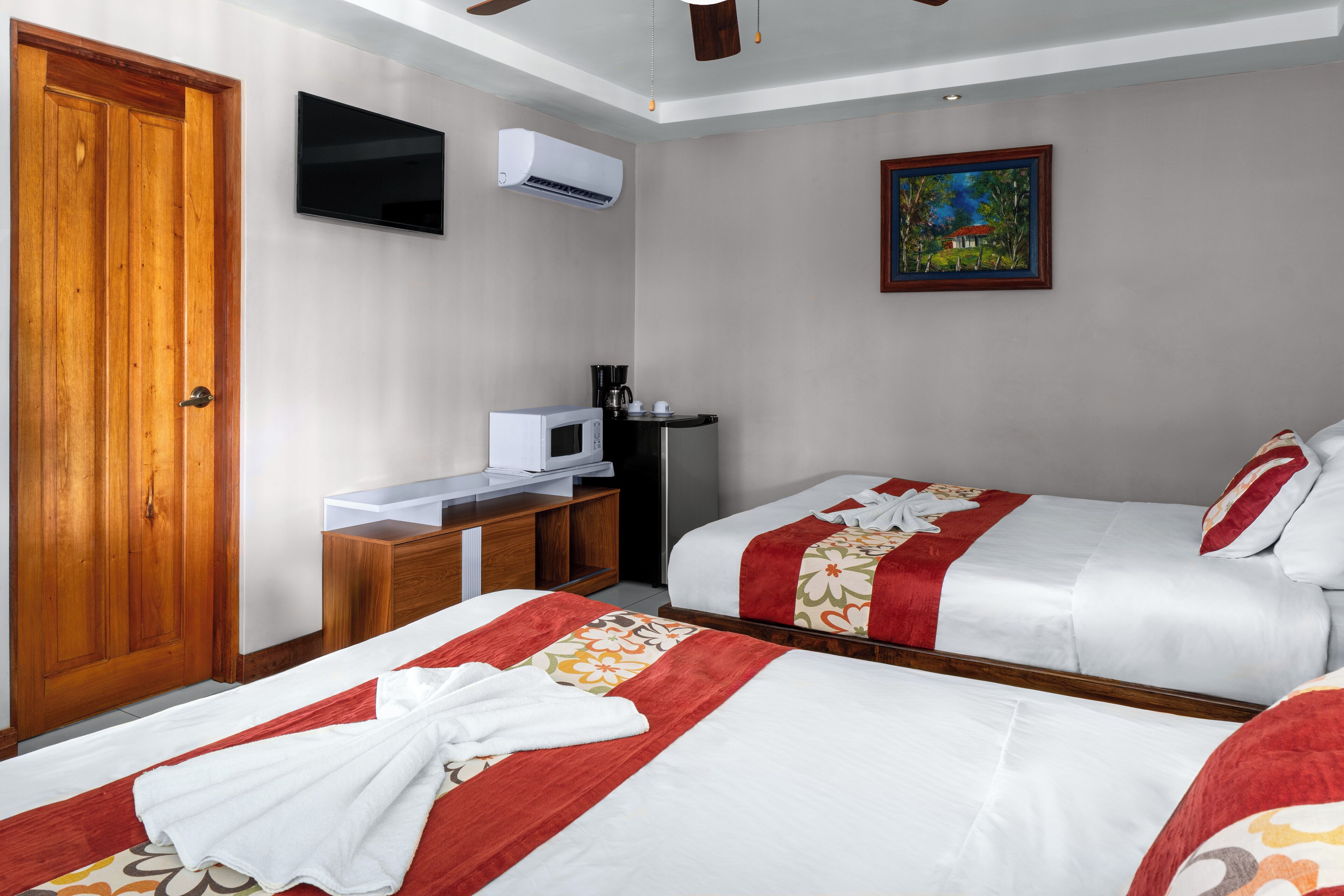 Habitación de hotel con dos camas, una puerta de madera, un televisor de pantalla plana, aire acondicionado, un cuadro y una kitchenette.