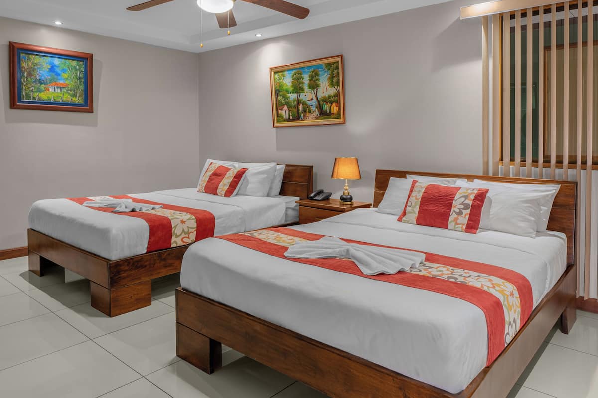 Quarto de hotel com duas camas de madeira, roupa de cama branca e detalhes em vermelho. Há duas pinturas nas paredes, um ventilador de teto e uma janela com persianas.