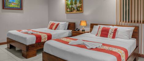 Habitación de hotel con dos camas de madera, ropa blanca y detalles en rojo. Hay dos cuadros en las paredes, un ventilador de techo y una ventana con persianas.
