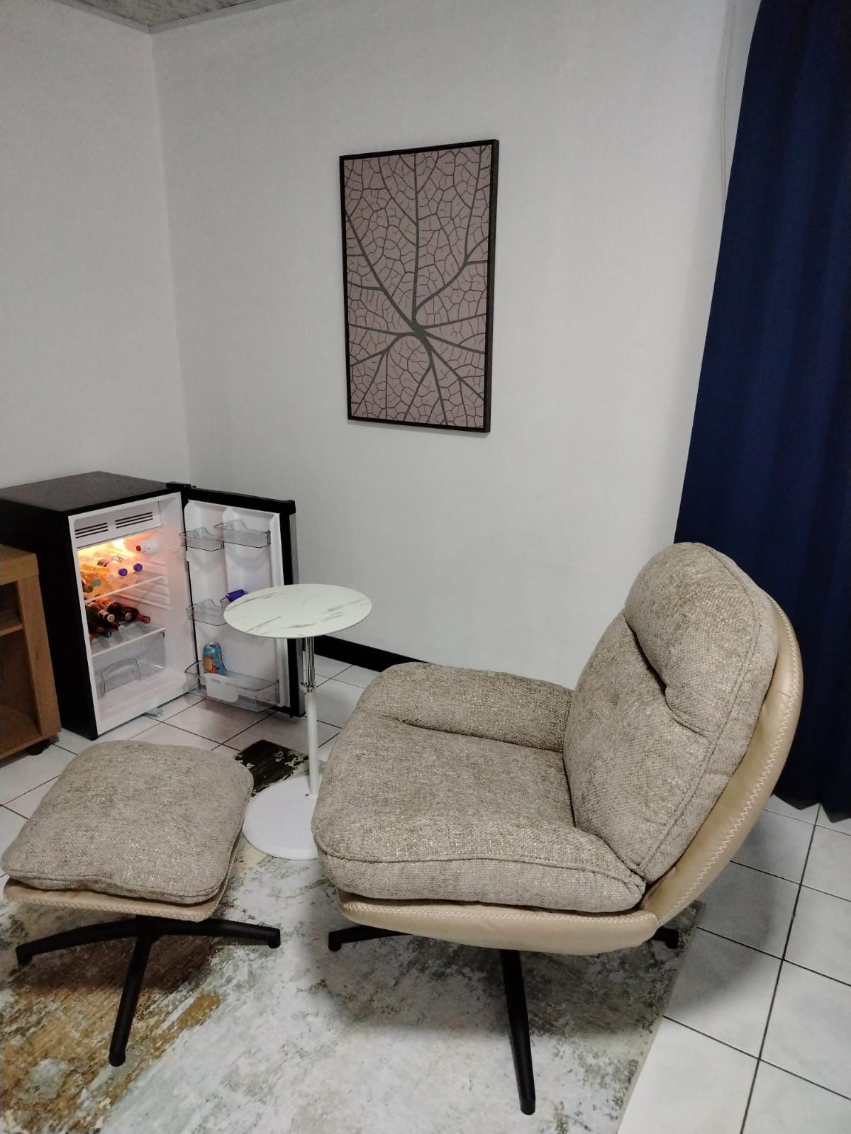 A living room with a sofa and ottoman, a small table, a refrigerator, and a wall-mounted abstract art piece.