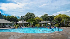 Una piscina con gente nadando y tomando el sol, rodeada de árboles y zonas de sombra.