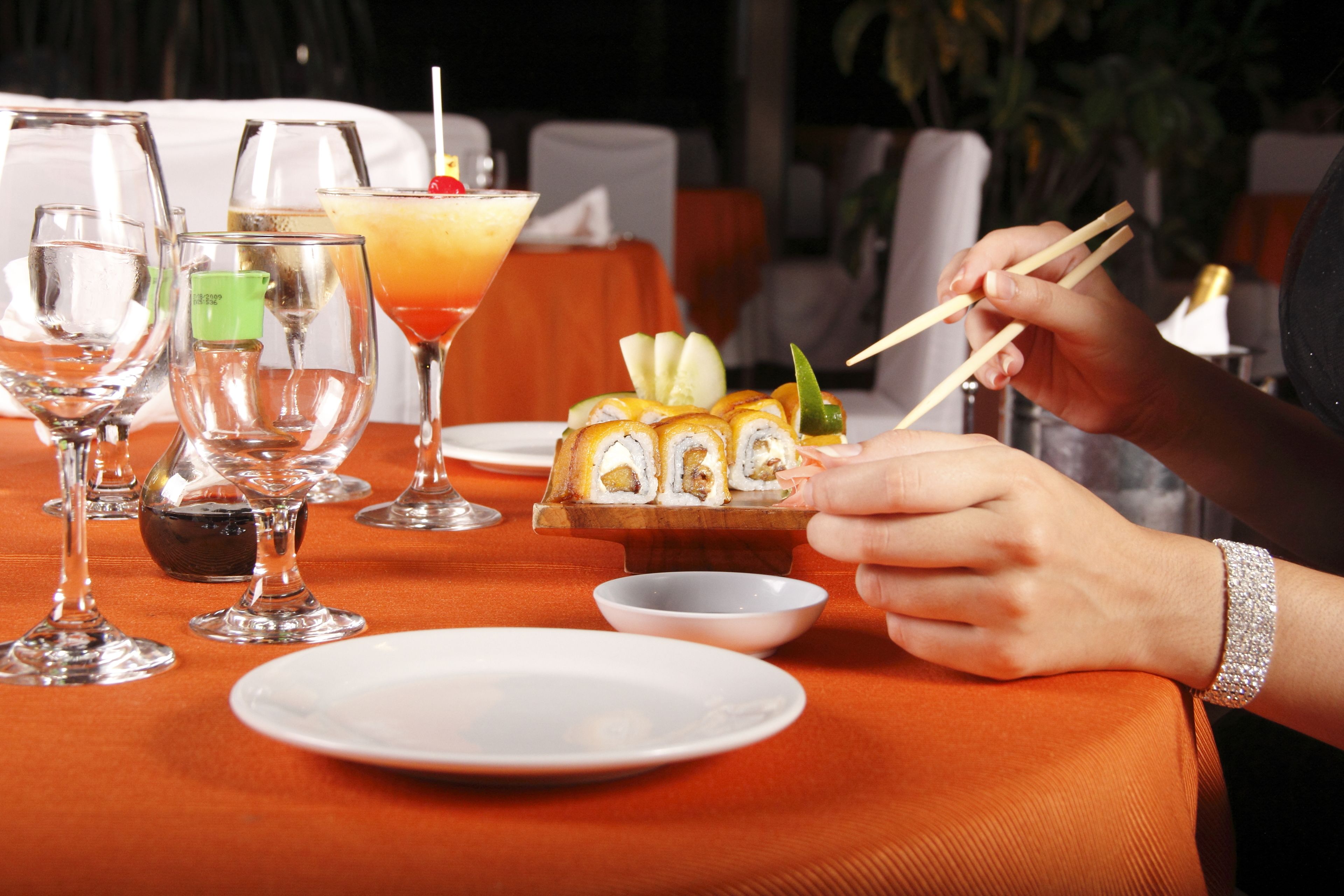 A dining table with various glasses, a cocktail, sushi, and a person using chopsticks.