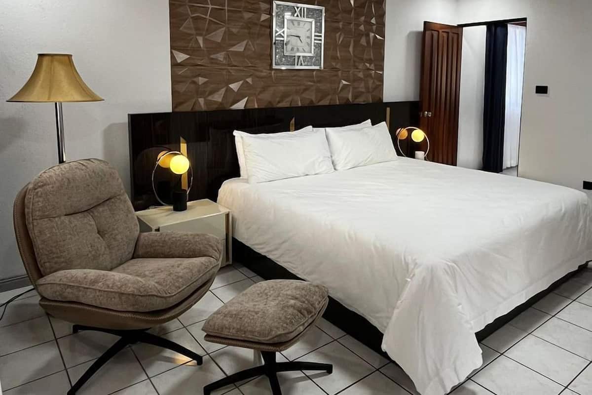 Quarto de hotel com cama, poltrona, escrivaninha, luminária e relógio de parede.