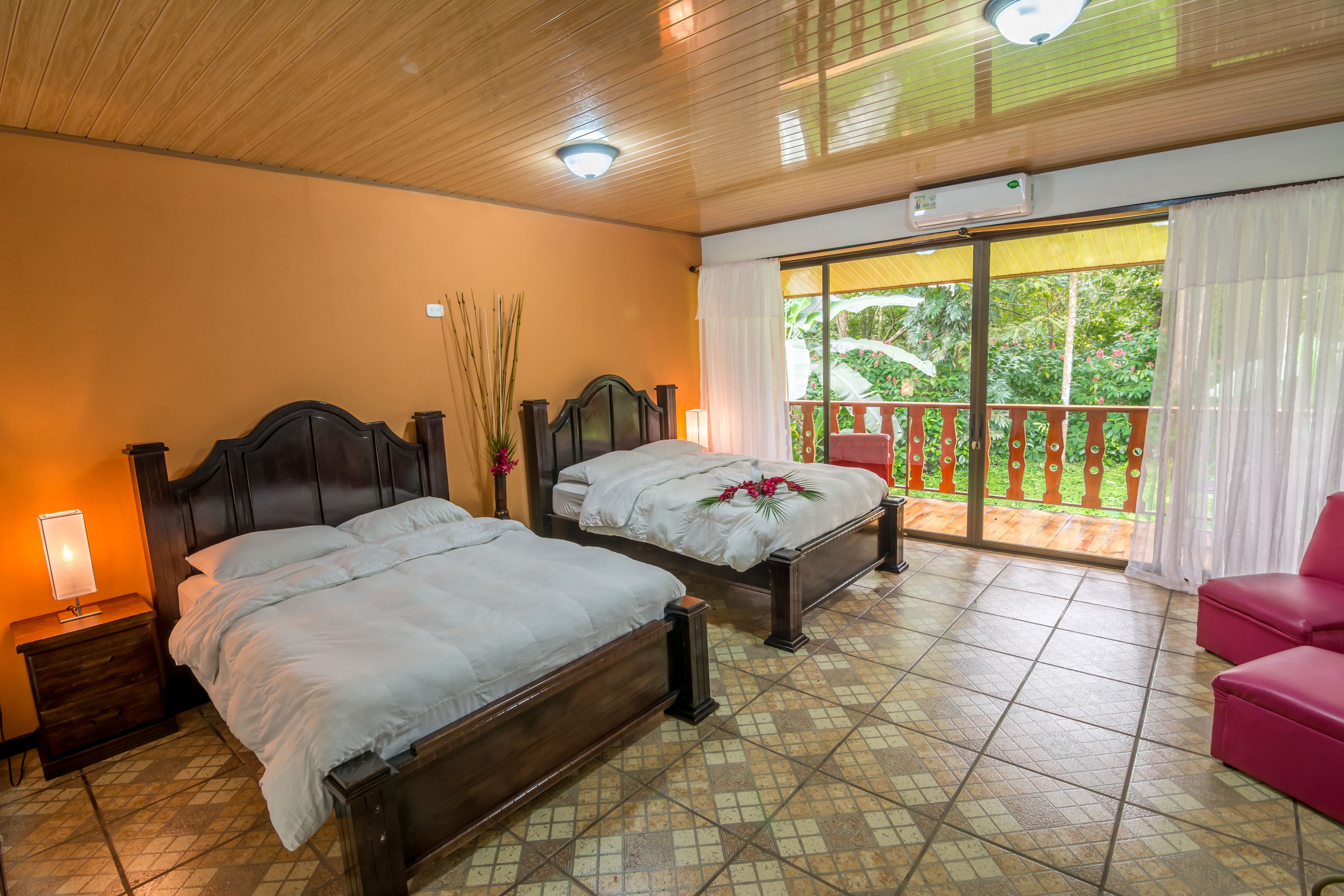 Une chambre d’hôtel avec deux lits en bois, une table de chevet avec une lampe, un balcon avec une chaise rouge et une vue sur la verdure environnante.