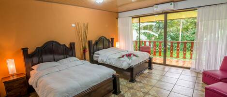 Une chambre d’hôtel avec deux lits en bois, une table de chevet avec une lampe, un balcon avec une chaise rouge et une vue sur la verdure environnante.