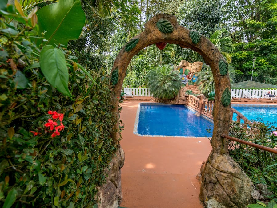 An outdoor pool area with a natural archway, surrounded by lush greenery and a white fence.