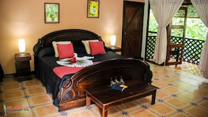 Une chambre à coucher avec un lit, des lampes de chevet, une tête de lit en bois, une table de chevet, un coussin décoratif, une lampe sur la table de chevet, une petite table avec un livre, et une vue sur la verdure à travers une fenêtre avec des rideaux.