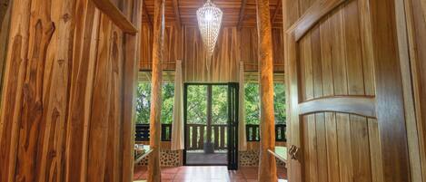 A wooden corridor with a chandelier, wooden doors, and a view of greenery outside.