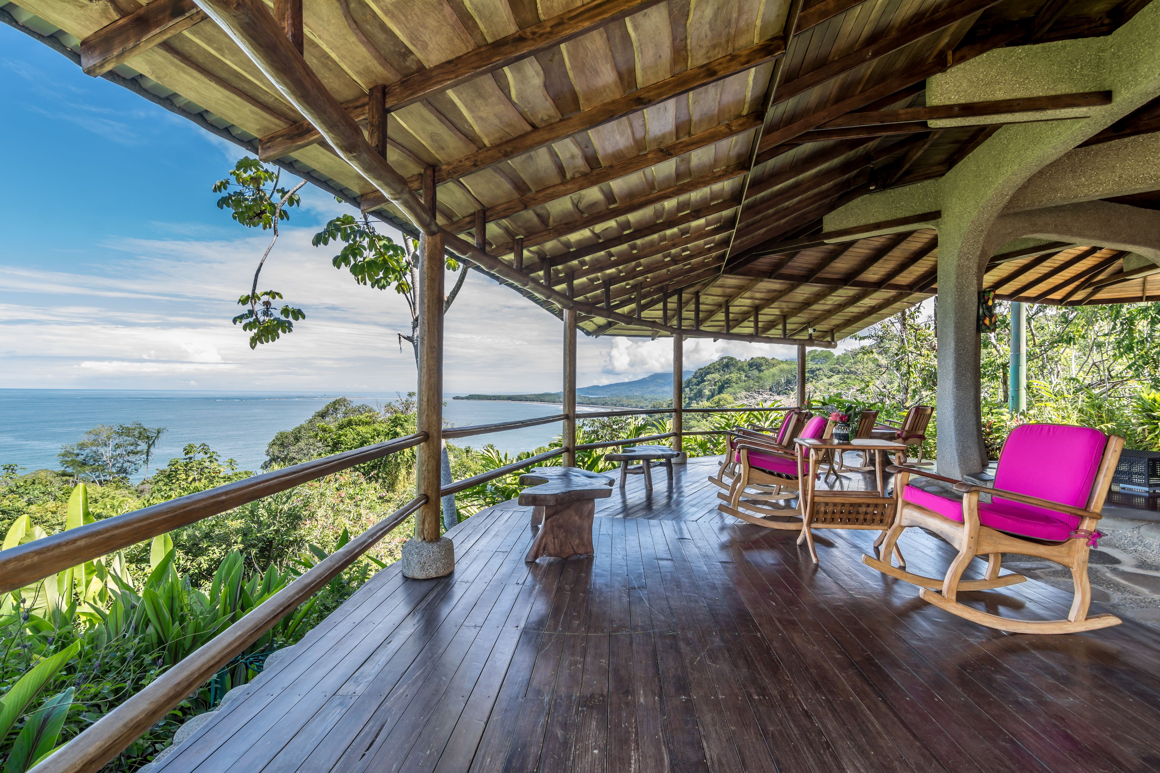 Terraza de madera al aire libre con vista al océano, sillas mecedoras de madera y una mesa.