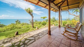 A covered outdoor area with a view of the ocean, a wooden rocking chair, and a stone pathway.