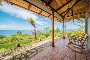 A covered outdoor area with a view of the ocean, a wooden rocking chair, and a stone pathway.